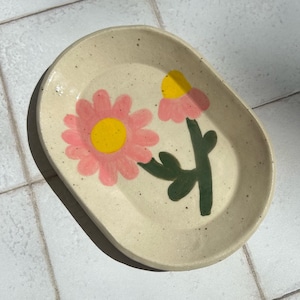 May include: Oval ceramic dish with a speckled, off-white base. The dish is decorated with two pink flowers with yellow centres and green stems. The flowers are hand-painted, adding a unique touch to the piece.