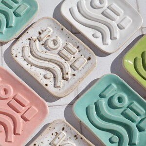 May include: A collection of rectangular ceramic soap dishes in various colours including pink, white, light green, and teal. Each dish features a raised relief design consisting of two wavy lines, a circle, and two rectangular bars.