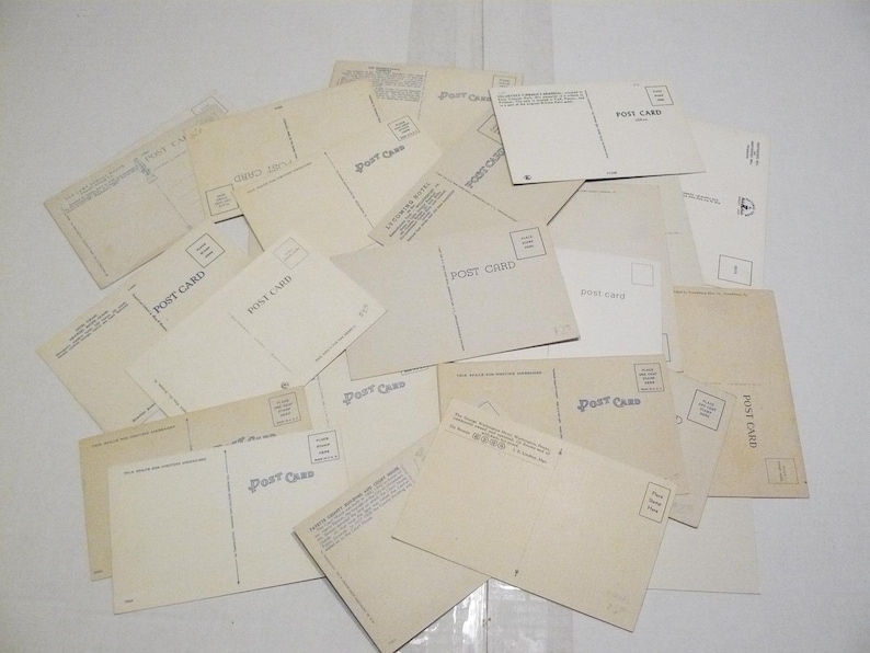 May include: A collection of vintage postcards, with the backs facing up, scattered across a surface. The postcards are primarily cream-colored with the words "POST CARD" printed on them. Some cards have a stamp box.