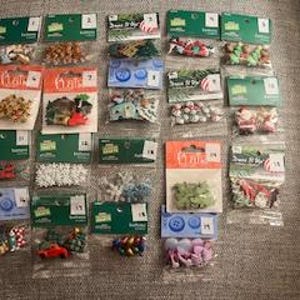 May include: A collection of small, packaged Christmas-themed craft embellishments. The packages contain various items like beads, buttons, and miniature figurines in festive colors such as red, green, and gold. The items are arranged on a textured, neutral-toned surface.