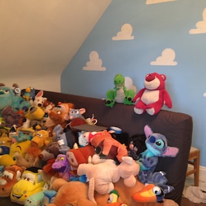 Puede incluir: Una colecci&oacute;n de peluches, incluyendo personajes de Toy Story, Monsters Inc., Lilo & Stitch y Buscando a Nemo, est&aacute;n apilados en una cama con una pared azul con recortes de nubes blancas.