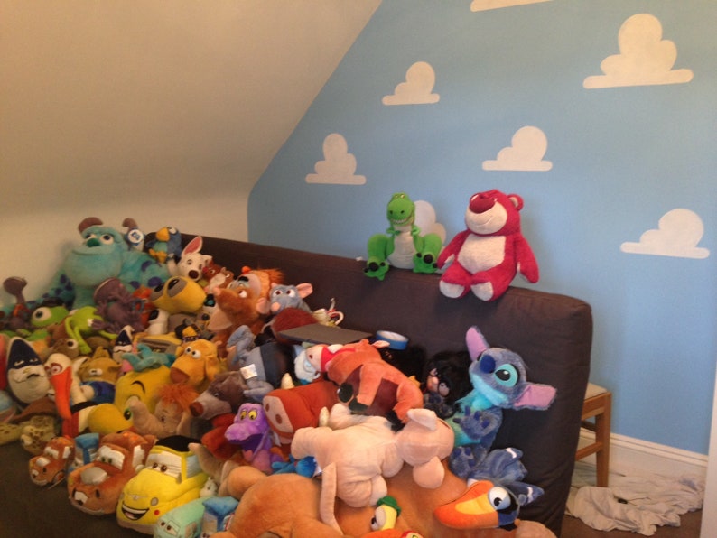 Puede incluir: Una colecci&oacute;n de peluches, incluyendo personajes de Toy Story, Monsters Inc., Lilo & Stitch y Buscando a Nemo, est&aacute;n apilados en una cama con una pared azul con recortes de nubes blancas.
