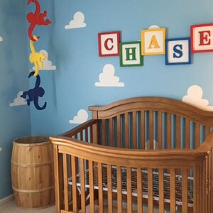 Puede incluir: Una habitaci&oacute;n de beb&eacute; con una cuna de madera, un barril de madera y un m&oacute;vil con tres monos colgando del techo. Las paredes est&aacute;n pintadas de azul y tienen nubes blancas y el nombre "CHASE" escrito en bloques de colores.