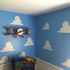 Puede incluir: Una pared azul con recortes de nubes blancas y una estanter&iacute;a de avi&oacute;n de madera roja con una h&eacute;lice blanca. La estanter&iacute;a est&aacute; montada en la pared.