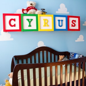 Puede incluir: Una habitaci&oacute;n de beb&eacute; decorada con un mural de pared de nubes azules y blancas. El nombre "CYRUS" est&aacute; escrito en bloques de madera coloridos sobre una cuna marr&oacute;n. Un oso de peluche y un robot de juguete est&aacute;n en el estante sobre los bloques.