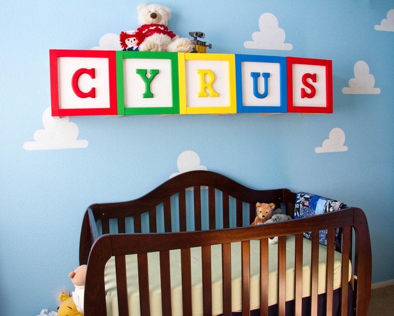Puede incluir: Una habitaci&oacute;n de beb&eacute; decorada con un mural de pared de nubes azules y blancas. El nombre "CYRUS" est&aacute; escrito en bloques de madera coloridos sobre una cuna marr&oacute;n. Un oso de peluche y un robot de juguete est&aacute;n en el estante sobre los bloques.