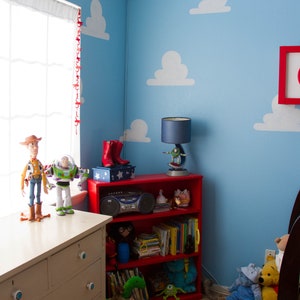 Puede incluir: Una estanter&iacute;a roja con juguetes y libros, una c&oacute;moda blanca con pomos blancos y una pared azul con nubes blancas pintadas en ella. La pared tambi&eacute;n tiene una cadena de luces navide&ntilde;as y una l&aacute;mpara roja, blanca y azul.