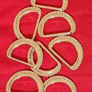 Op de afbeelding: Een groep van acht gouden metalen D-ringen met een gevlochten ontwerp. De ringen liggen op een rode ondergrond.