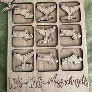 May include: A wooden tic-tac-toe game with a Massachusetts state outline and whale tail pieces. The game is engraved with the words "Tic-Tac-Toe Massachusetts".