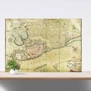Op de afbeelding: Een antieke kaart van Riga, Letland, met een kompasroos en de tekst "Plan der Stadt Festung Riga 1764". De kaart is gedrukt op verouderd papier met een gelige tint en is ingelijst in een eenvoudig houten frame.