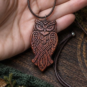 Può includere: Un ciondolo a forma di gufo in legno marrone scuro con intricati motivi a intreccio intagliati e un simbolo triskelion centrale. Il ciondolo è appeso a una sottile collana in cordino intrecciato marrone scuro con chiusura a perla scorrevole.