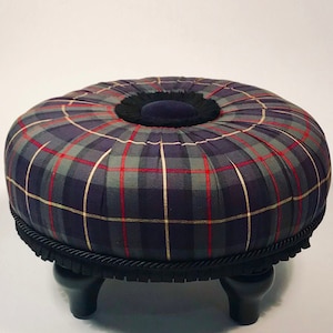 May include: A round, tufted ottoman with a plaid fabric cover in shades of blue, green, red, and cream. The ottoman has a black velvet center and a black braided trim around the base. It sits on four black wooden legs.