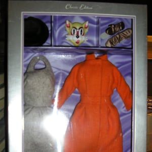 May include: A silver box with a clear plastic window showing a doll outfit. The outfit includes a red dress, a gray coat, and a pair of black shoes. The box also includes a black purse and a cat head. The box has the text "Classic Edition" and "Paramount Pictures Audrey Hepburn Collection" on it.