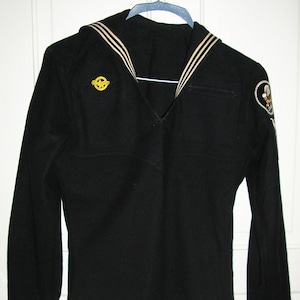 May include: A black long-sleeved shirt with a white sailor collar and two white stripes on each side. The shirt has a zipper on the right side and a pocket on the left side. There are two patches on the shirt, one on the left chest and one on the right sleeve.