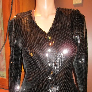 May include: A black sequined top with a v-neck and long sleeves. The top has a button closure and is paired with a green and black patterned skirt.