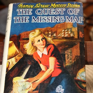 May include: Vintage Nancy Drew Mystery Stories book titled "The Quest of the Missing Map." The cover features an illustration of a woman in a red shirt and blue skirt, holding a map. The book has a blue background with the title and author's name.
