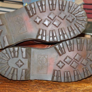 Op de afbeelding: Close-up van de zolen van twee bruine leren laarzen. De zolen zijn gemaakt van een getextureerd, donkerbruin rubber met een ruitpatroon. De woorden "Made in England" zijn zichtbaar.