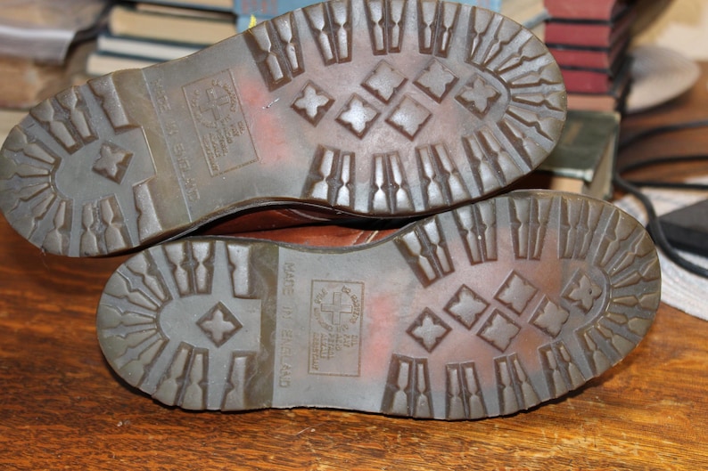 Op de afbeelding: Close-up van de zolen van twee bruine leren laarzen. De zolen zijn gemaakt van een getextureerd, donkerbruin rubber met een ruitpatroon. De woorden "Made in England" zijn zichtbaar.
