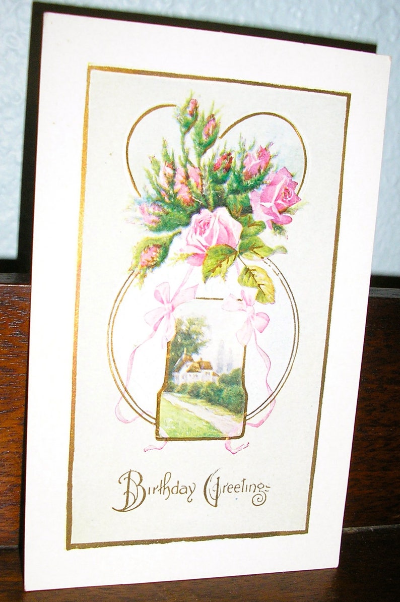 May include: A vintage postcard with a gold border and the text "Birthday Greetings". The postcard features a bouquet of pink roses in a vase with a landscape scene. The roses are arranged in a heart shape.