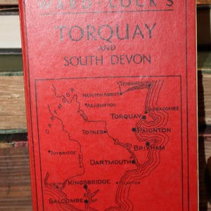 May include: A vintage red travel guide titled "Ward Lock's Torquay and South Devon." The cover features a map of the region with place names like Torquay, Paignton, and Dartmouth. The book is surrounded by other books.