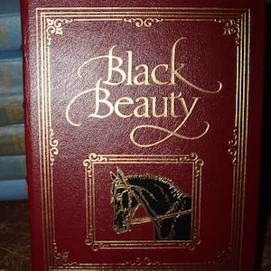 K&ouml;nnte beinhalten: Ein Hardcover-Buch mit dem Titel "Black Beauty" mit burgunderfarbenem Einband und goldener Schrift. Das Cover zeigt eine goldgerahmte Illustration eines Pferdekopfes und dekorative goldene R&auml;nder.