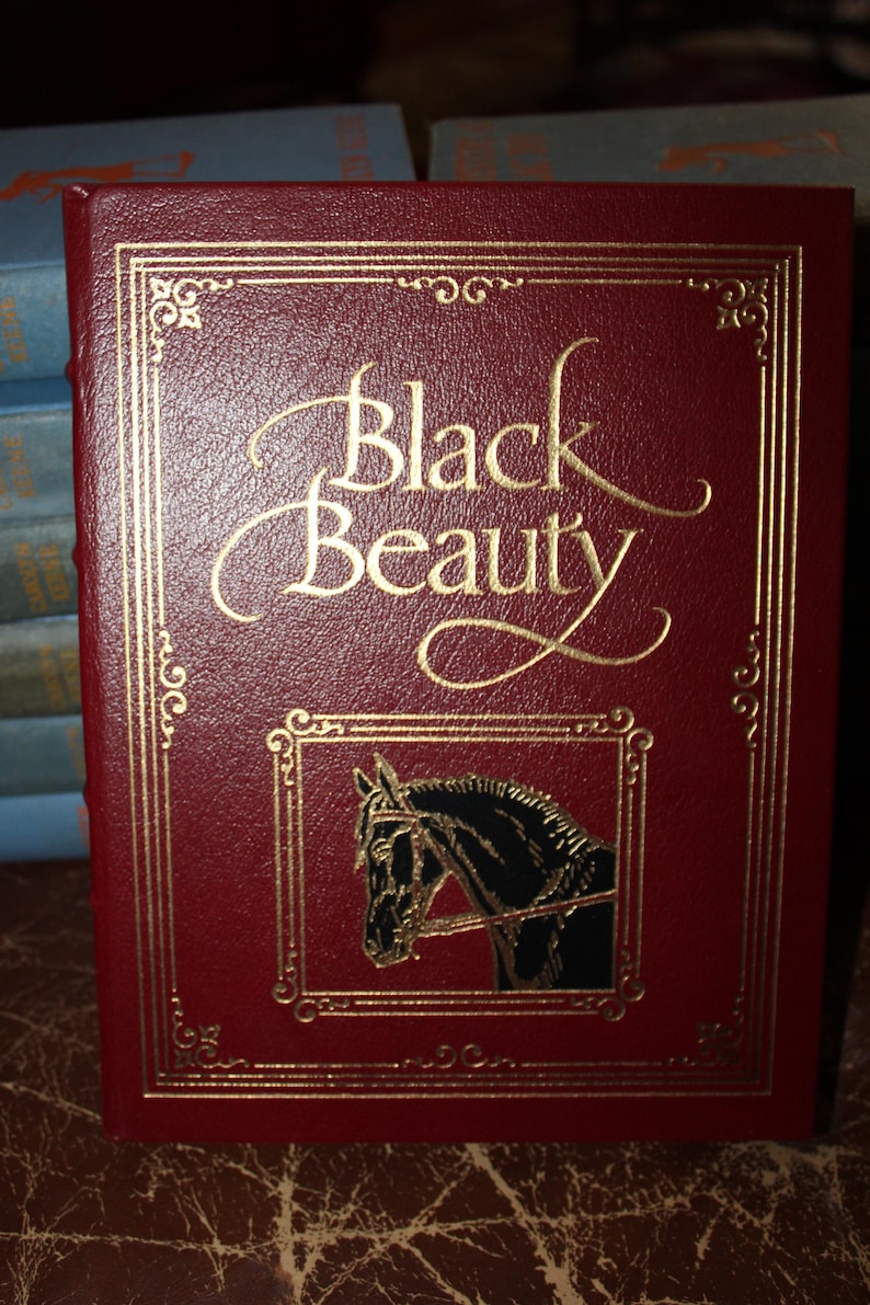 K&ouml;nnte beinhalten: Ein Hardcover-Buch mit dem Titel "Black Beauty" mit burgunderfarbenem Einband und goldener Schrift. Das Cover zeigt eine goldgerahmte Illustration eines Pferdekopfes und dekorative goldene R&auml;nder.