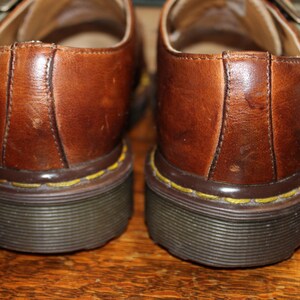 Op de afbeelding: Close-up van twee bruine leren schoenen met een gespsluiting. De schoenen hebben een donkere zool met gele stiksels en een bruine rand. De leren bovenzijde vertoont slijtage.