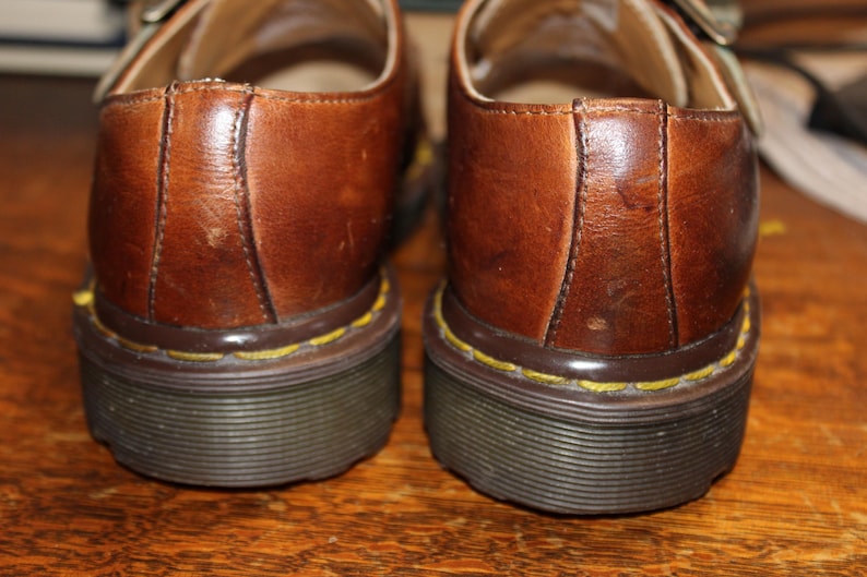 Op de afbeelding: Close-up van twee bruine leren schoenen met een gespsluiting. De schoenen hebben een donkere zool met gele stiksels en een bruine rand. De leren bovenzijde vertoont slijtage.