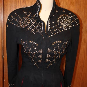 May include: Black jacket with gold-colored studs and rhinestones. The jacket has a high collar, long sleeves, and a zipper closure. The jacket is displayed on a mannequin with a red skirt.