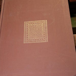 May include: A brown book with a gold square design on the cover. The square design is made up of a brick pattern. The book is sitting on a wooden surface.