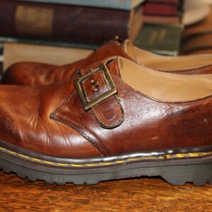 Op de afbeelding: Een paar bruine leren schoenen met een gespsluiting. De schoenen hebben een dikke, donkere zool met gele stiksels langs de rand. Het leer heeft een versleten uiterlijk. De schoenen zijn ongeveer 25 cm lang.