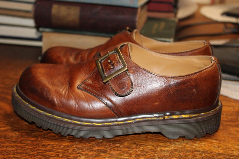 Op de afbeelding: Een paar bruine leren schoenen met een gespsluiting. De schoenen hebben een dikke, donkere zool met gele stiksels langs de rand. Het leer heeft een versleten uiterlijk. De schoenen zijn ongeveer 25 cm lang.