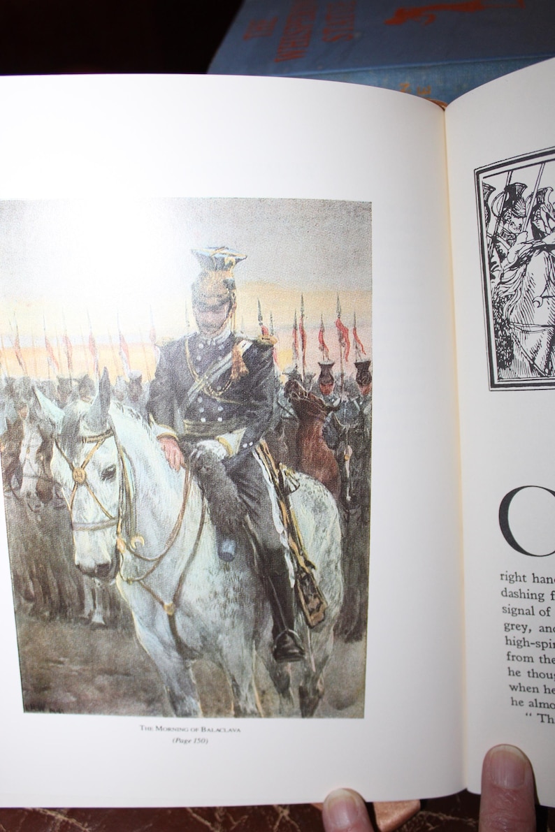K&ouml;nnte beinhalten: Buchillustration zeigt einen Mann in dunkler Uniform auf einem wei&szlig;en Pferd, der Soldaten anf&uuml;hrt. Das Kunstwerk verwendet ged&auml;mpfte Farben, mit roten Fahnen im Hintergrund. Der Titel "The Morning of Balaclava" befindet sich am unteren Bildrand.