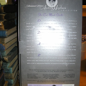 May include: A silver box with a photo of Audrey Hepburn wearing a black dress and a cat mask. The box is for a collectible doll and features the text "Paramount Pictures Audrey Hepburn Collection The Cat Mask Outfit".