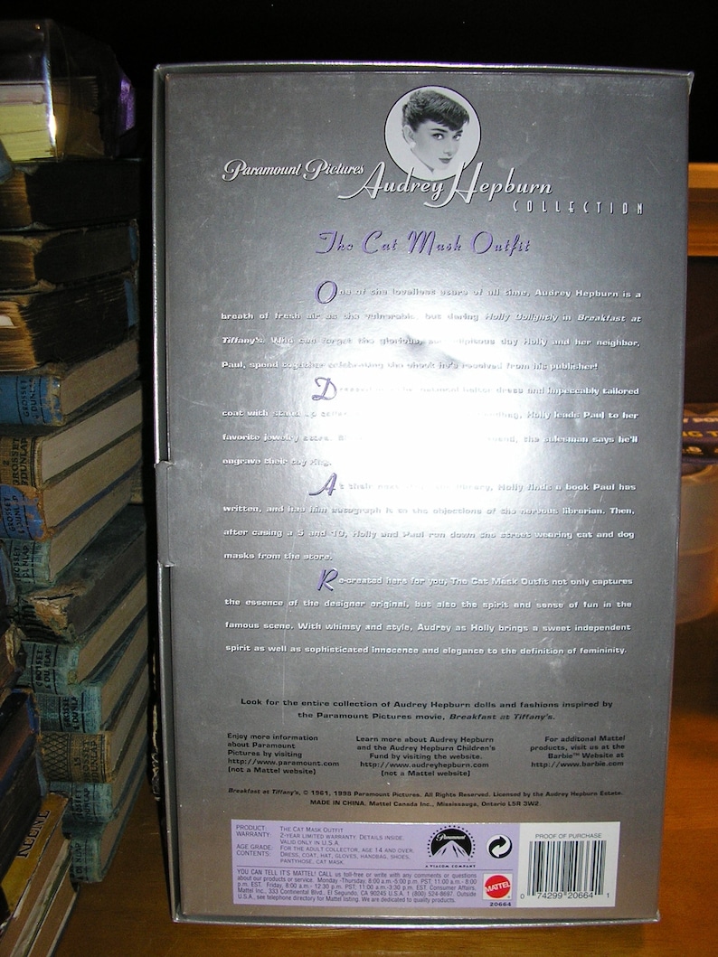May include: A silver box with a photo of Audrey Hepburn wearing a black dress and a cat mask. The box is for a collectible doll and features the text "Paramount Pictures Audrey Hepburn Collection The Cat Mask Outfit".