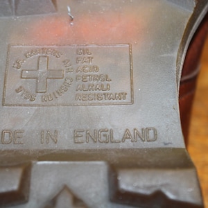 Op de afbeelding: Close-up van de zool van een bruine laars. De zool is voorzien van het Dr. Martens-logo, een kruis in een cirkel, en de tekst "OIL FAT ACID PETROL ALKALI RESISTANT". De woorden "MADE IN ENGLAND" zijn ook zichtbaar.