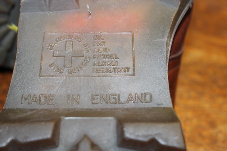 Op de afbeelding: Close-up van de zool van een bruine laars. De zool is voorzien van het Dr. Martens-logo, een kruis in een cirkel, en de tekst "OIL FAT ACID PETROL ALKALI RESISTANT". De woorden "MADE IN ENGLAND" zijn ook zichtbaar.