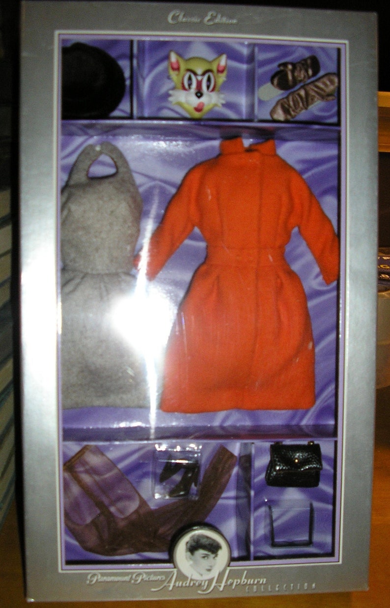 May include: A silver box with a clear plastic window showing a doll outfit. The outfit includes a red dress, a gray coat, and a pair of black shoes. The box also includes a black purse and a cat head. The box has the text "Classic Edition" and "Paramount Pictures Audrey Hepburn Collection" on it.