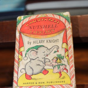 May include: A vintage children's book titled "Christmas Nutshell Library" by Hilary Knight. The cover features an illustration of an elephant and a girl. The book has a red and green color scheme with the publisher's name at the bottom.