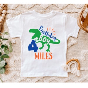 Peut inclure: T-shirt blanc avec un motif de dinosaure vert, le chiffre 4 bleu et le texte "Birthday Boy Miles".