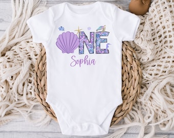 May include: White baby onesie with a purple seashell and the word "ONE" in purple with a mermaid scale pattern. The name "Sophia" is written below the word "ONE".