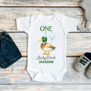 May include: A white short-sleeved baby bodysuit featuring a graphic of a mallard duck wearing a blue party hat and bow tie with a balloon. The text reads ONE, Lucky Duck, and JACKSON in green and grey lettering, with three snaps at the bottom.