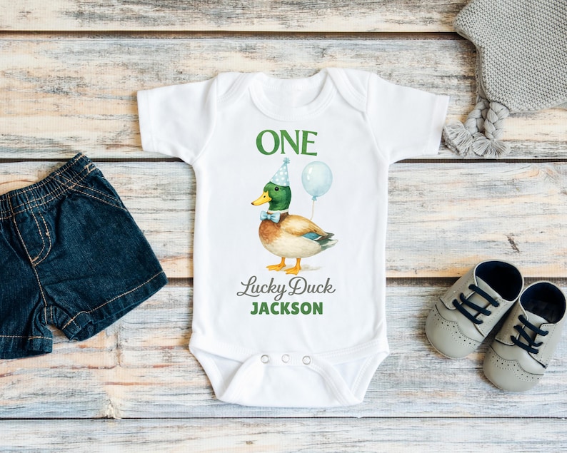 May include: A white short-sleeved baby bodysuit featuring a graphic of a mallard duck wearing a blue party hat and bow tie with a balloon. The text reads ONE, Lucky Duck, and JACKSON in green and grey lettering, with three snaps at the bottom.