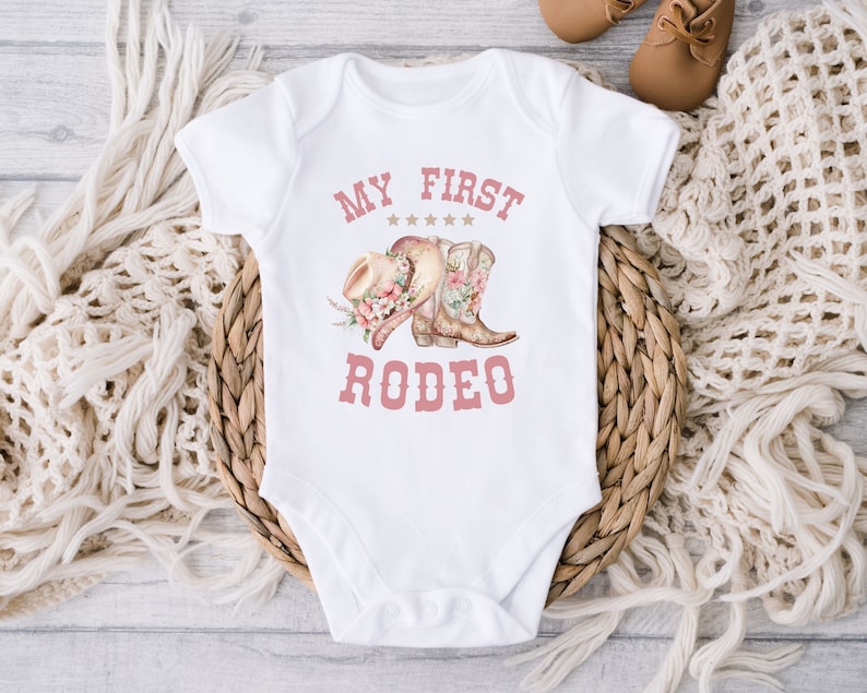 May include: White baby onesie with pink text that reads "My First Rodeo". The onesie features a pink cowboy hat and boots with floral accents.