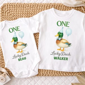 May include: A white baby bodysuit and a white t-shirt, both featuring a graphic of a mallard duck wearing a blue party hat and bow tie with a light blue balloon. The text reads ONE, Lucky Duck, and the names BEAU and WALKER in green and grey.