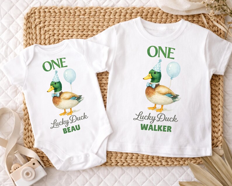 May include: A white baby bodysuit and a white t-shirt, both featuring a graphic of a mallard duck wearing a blue party hat and bow tie with a light blue balloon. The text reads ONE, Lucky Duck, and the names BEAU and WALKER in green and grey.