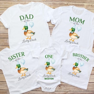 May include: A collection of five white t-shirts and one white bodysuit featuring a mallard duck wearing a blue party hat and bow tie, a light blue balloon, and text including DAD, MOM, SISTER, BROTHER, ONE, and the name WESTON in green.