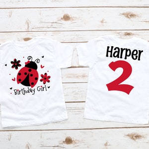 May include: Two white t-shirts with a ladybug design. The front shirt has a ladybug with black spots and red wings, surrounded by red hearts and black flowers. The text "Birthday Girl" is below the ladybug. The back shirt has the name "Harper" and the number "2" in red.