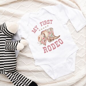 May include: White long-sleeved baby onesie with pink text that reads "My First Rodeo" and a graphic of pink cowboy boots and a hat with flowers.
