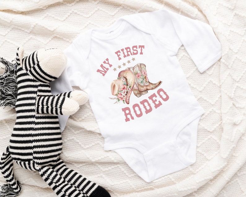 May include: White long-sleeved baby onesie with pink text that reads "My First Rodeo" and a graphic of pink cowboy boots and a hat with flowers.